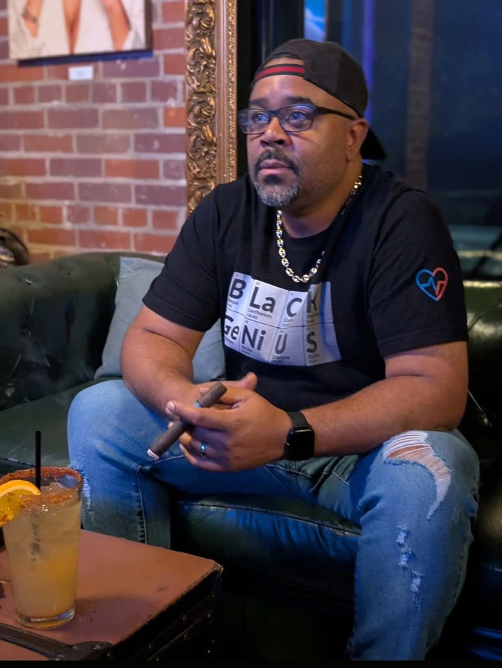 Man sitting on a couch next to a drink and a cigar, with a brick wall and a framed picture in the background. Wearing Black Genius T Shirt