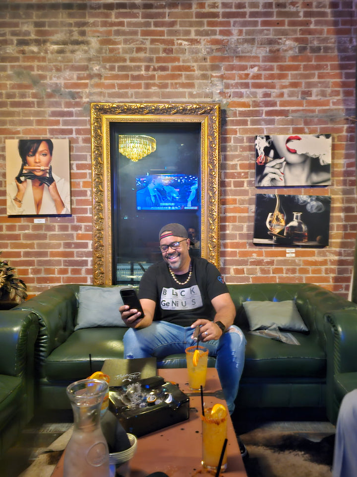 Man sitting on a green couch in a room with brick walls, framed pictures, and a TV. Wearing Black Genius T Shirt Smiling