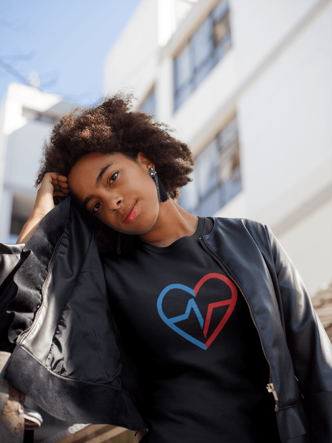 Black woman wearing a black Love Clan Signature Tee and leather jacket, standing outdoors in natural light with a modern building in the background.