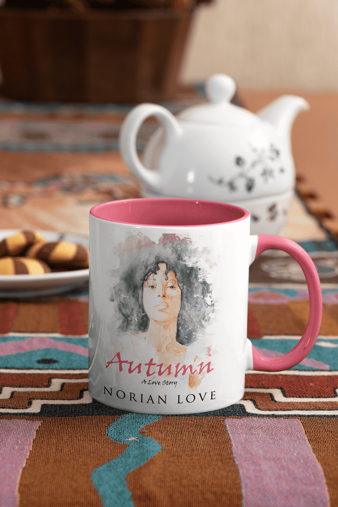 Autumn mug with pink interior beside a teapot and biscuits on a patterned table runner.