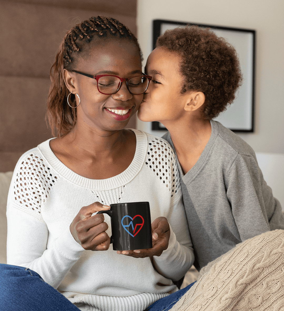 Black ceramic mug with Love Clan heart design held by a smiling Black mother as her son kisses her cheek.
