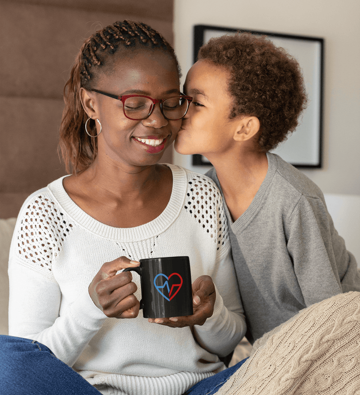 Black ceramic mug with Love Clan heart design held by a smiling Black mother as her son kisses her cheek.