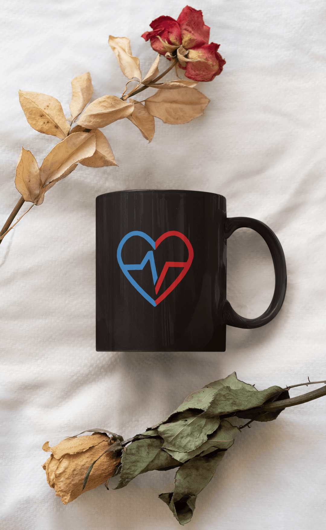 Black mug with Love Clan heart design displayed between dried roses on a white fabric background.