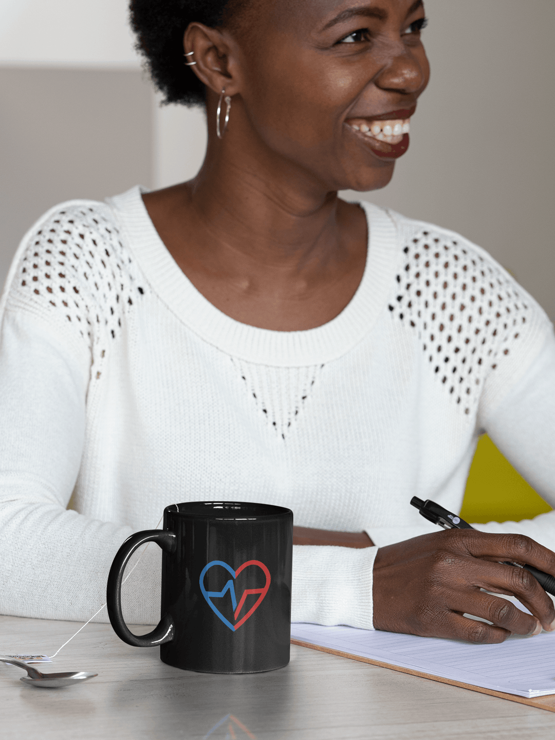 Black coffee mug with Love Clan heart design on a table next to a smiling Black woman writing in a notebook