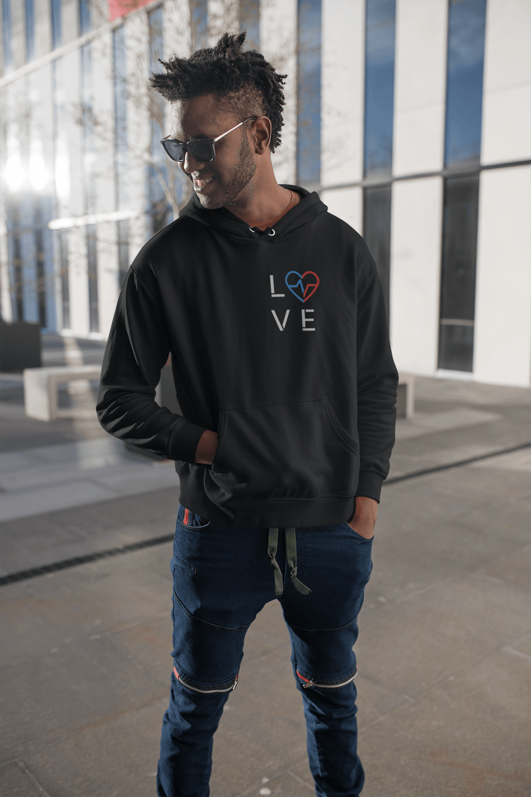Black man wearing the Love Season Hoodie featuring the red and blue Love Clan emblem on a black hoodie, standing outdoors in a modern urban setting.