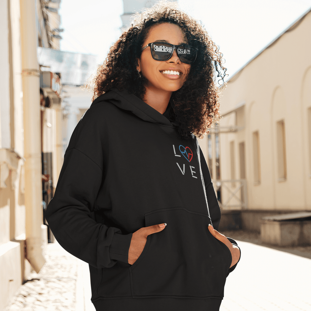 Black woman wearing the Love Season Hoodie with the red and blue Love Clan emblem, standing outdoors in natural light in a casual streetwear setting.