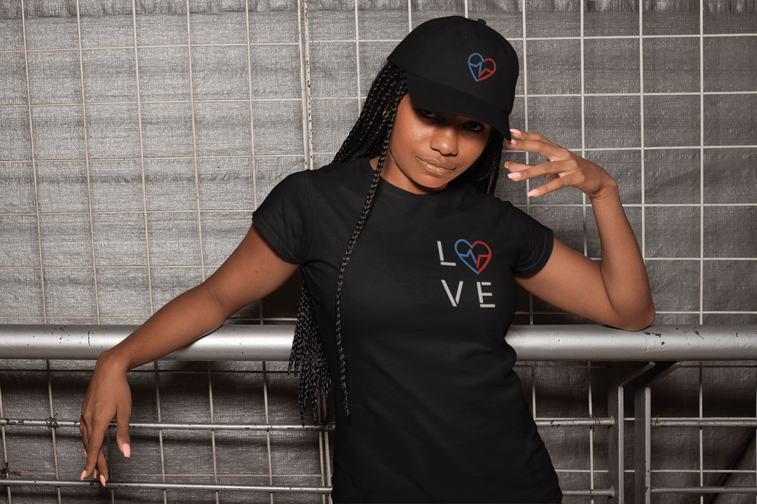 Black woman wearing a black Love Season Tee with the red and blue Love Clan emblem and minimalist LOVE design, paired with a matching embroidered hat, posing against an industrial metal backdrop.