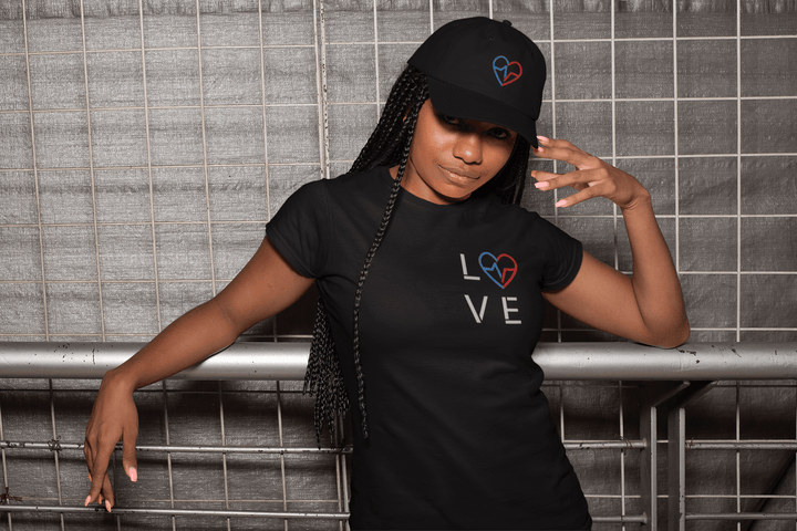 Black woman wearing a black Love Season Tee with the red and blue Love Clan emblem and minimalist LOVE design, paired with a matching embroidered hat, posing against an industrial metal backdrop.
